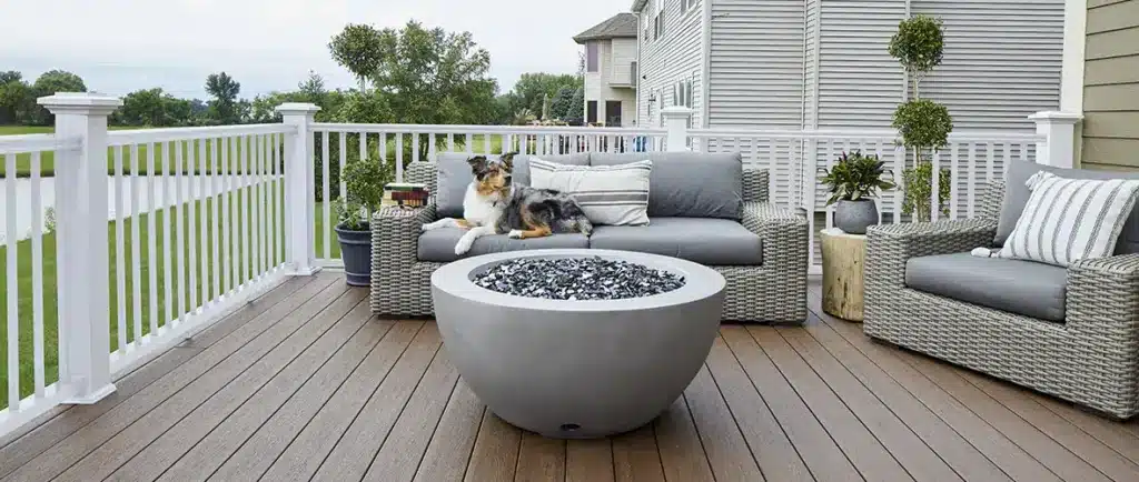 A dog lounges on outdoor furniture in front of a firepit on a deck.